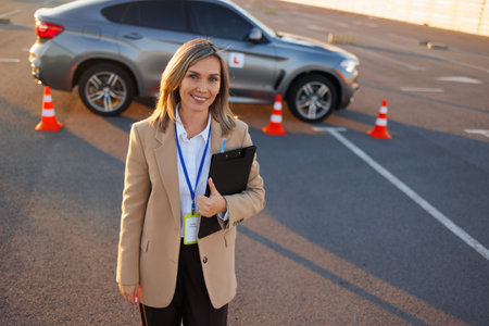 Smiling driving instructor holding clipboard near learner car on test siteの写真素材