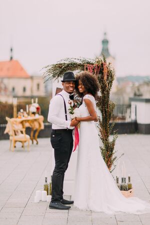 Cheerful black bride and groom holding hands on the wedding ceremony on rooftop.の写真素材