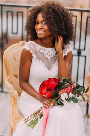 Happy young black bride smiling with bouquet of red flowers and sitting on vintage terracotta chair.の写真素材