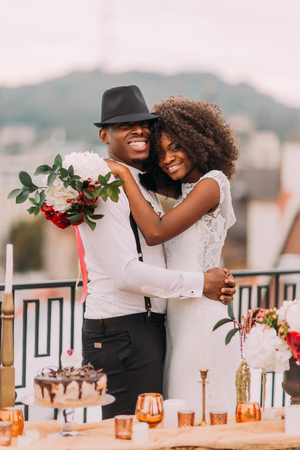 Happy black bride and stylish groom posing for camera on the terrace with cityscape on foreground.の写真素材