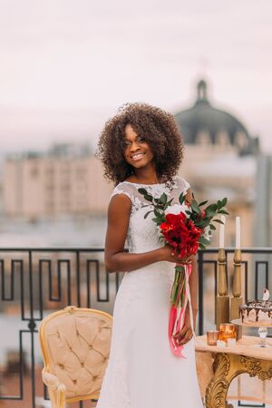 Beautiful african bride with wedding bouquet on the rooftop with ancient european architecture on background.の写真素材