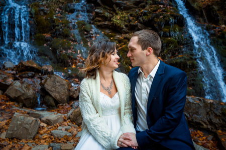 Wedding couple sitting on the stone. Waterfall background. Honeymoon in autumn mountains.の写真素材