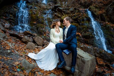 Wedding couple sitting on stone and lovingly look at each other. Waterfall background.の写真素材