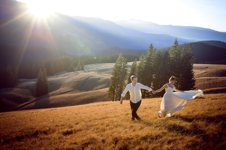 Beautiful wedding couple running and having fun on the field surrounded by mountains.の写真素材