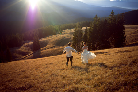 Happy wedding couple running and having fun on the field surrounded by mountains.の写真素材