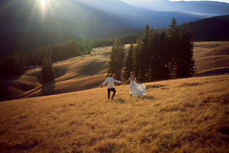 Happy wedding couple running and having fun on the field surrounded by mountains.の写真素材