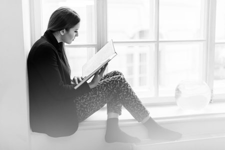 Beautiful stylish young girl reading a book on the windowsill. Black and white photo.