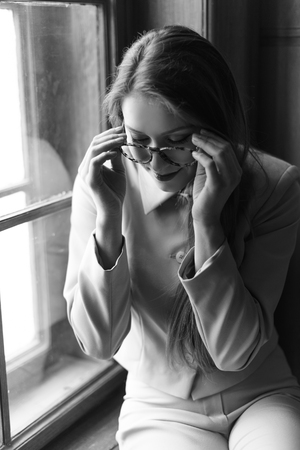 Portrait of young beautiful girl in glasses on windowsill. Black and white photo.の写真素材
