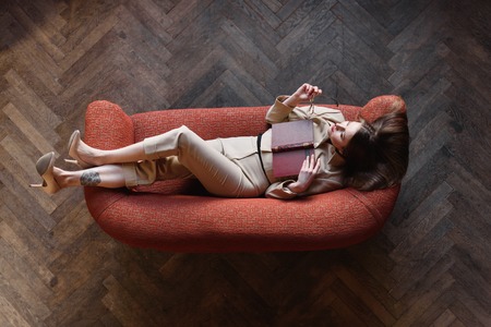 Stylish young girl in beige suit reads a book on the red sofa. Top view.の写真素材