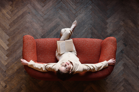 Stylish young girl in beige suit reads a book on the red sofa. Top view.の写真素材