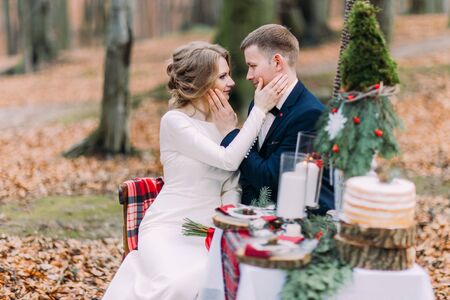 Charming wedding couple softly stroking face of each other at the holiday table in autumn forest.の写真素材