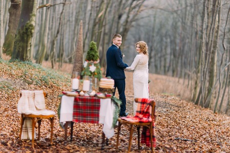 Happy young wedding couple walking and holding hands near the decorated table for Christmas holidays in autumn forestの写真素材