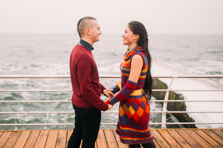 Bright stylish couple holding hands and lovingly look at each other on the berth. Sea background. Wonderful honeymoon.の写真素材