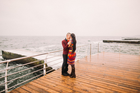 Young hipster bright couple in love tenderly holding hands and hugs at lonely beach on cloudy day. Sea background. Beautiful honeymoon.の写真素材