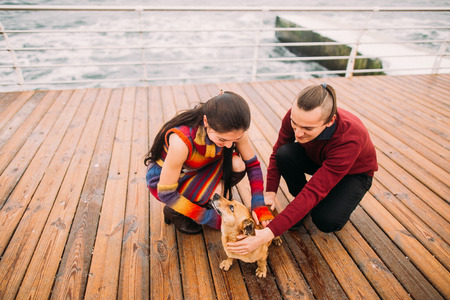 Young happy couple playing with dog on the rainy berth in autumn. Sea background.の写真素材