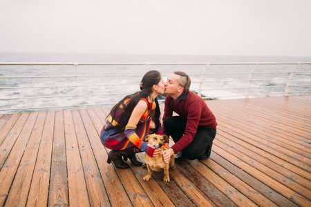 Young happy couple kissing each other and playing with dog on the rainy berth in autumn. Sea background.の写真素材