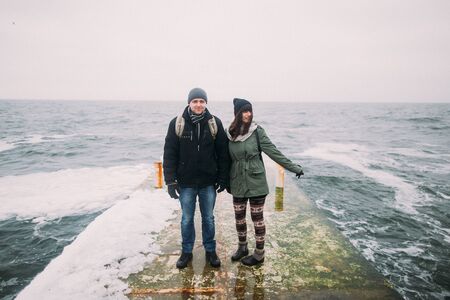 Happy stylish couple posing and holding hands on the old pier in winter vacation. Travel together concept. Sea background.の写真素材