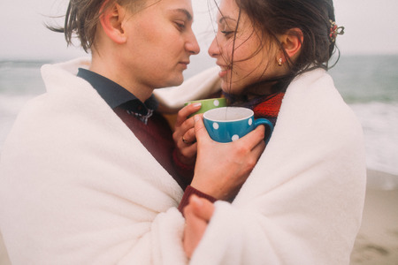 Beautiful happy young couple lovingly look at each other wrapped in white blanket with cute cups of tea. Winter beach on background.の写真素材