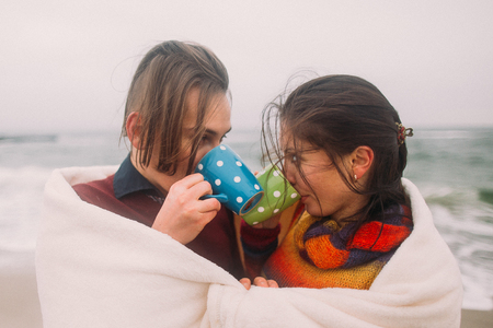 Beautiful happy young couple lovingly look at each other wrapped in white blanket and drinking tea from cute colorful cups. Winter beach on background.の写真素材