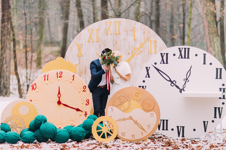 Happy wedding couple kissing at the big vintage clocks in  autumn forest. Creative wedding decorations.の写真素材
