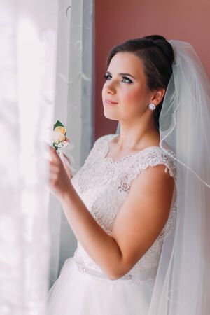 Beautiful brunet bride in white wedding dress standing in her bedroom near the window.の写真素材