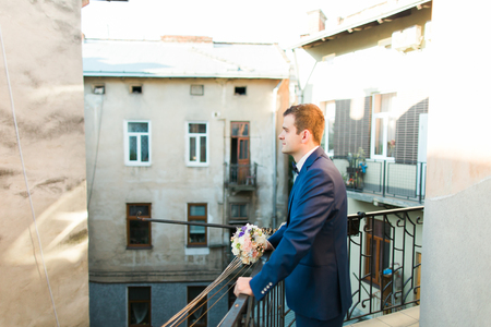 Cute stylish groom in blue suit and bow tie with a bouquet of roses standing at balcony looking towards.の写真素材