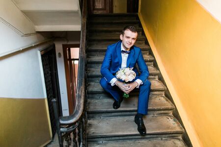 Young attractive smiling man in blue suit sitting on stairs with bouquet, indoor shot.の写真素材