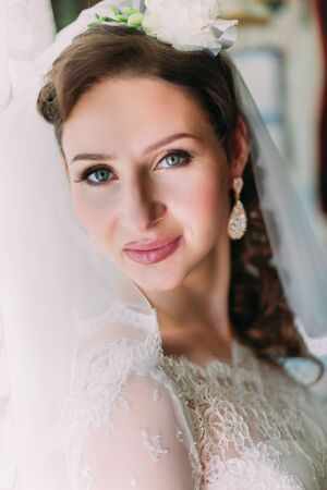 Close up portrait of happy smiling bride in luxury white dress and veil posing with pink rose near the window.の写真素材