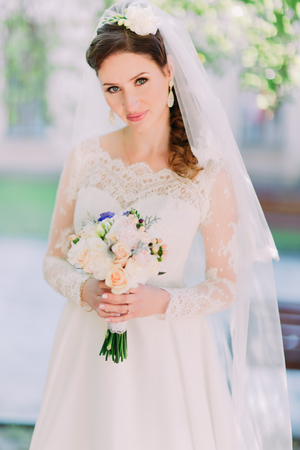 Cheerful young brunette woman with pink lips and a bouquet. Beauty portrait perfect makeup. Long elegant chic curly hair. Wedding day, white dress. On the street in the park.の写真素材