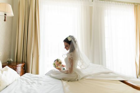 Happy stylish  bride under the veil in luxury white dress sitting near the windowの写真素材