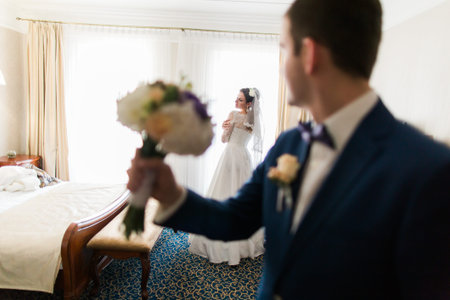 Handsome groom with bouquet posing at beautiful bride background in hotel room.の写真素材