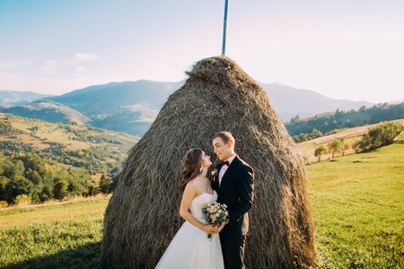 Beautiful young bride is tenderly embracing husband at rural haystacks summer field background.の写真素材