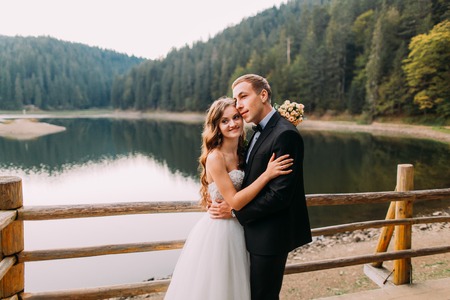 Elegant beautiful wedding couple embracing near wooden fence on lake background.の写真素材