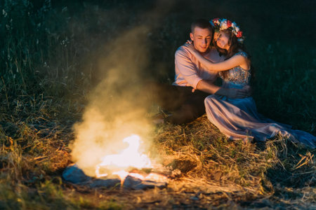 Young couple: girl in wreath and sitting holding hands face-to-face around the campfire at night