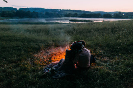 Happy young couple sitting back on plaid embracing by fire at night. Girl in wreath.