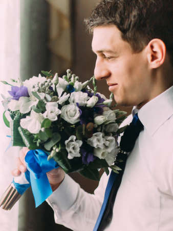 Stylish handsome dark haired groom sniffing a wedding bouquet standing near the window.の写真素材