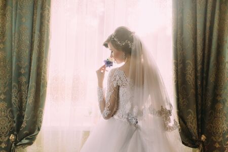 Silhouette of a beautiful bride in a traditional white wedding dress, stood by window holding buttonholeの写真素材
