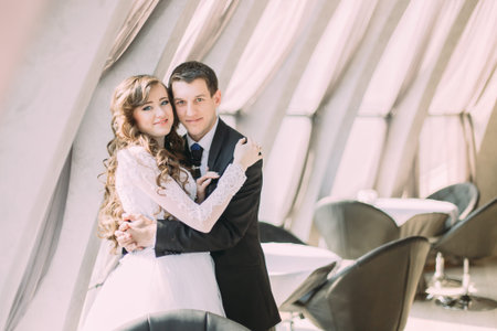Stylish bride and groom embracing looking at the camera near window in a room with white interior.