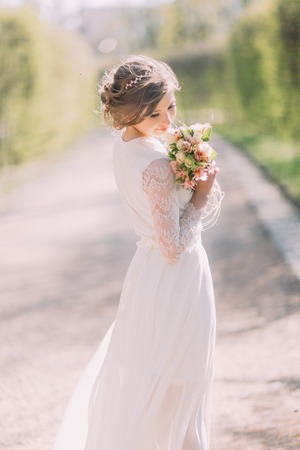 Back view of young blonde bride in white dress with bridal bouquet of spring flowers standing outdoor.の写真素材