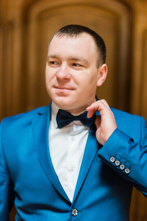 Handsome smiling man in blue suit fixin his bow tie in rich wooden interior.の写真素材