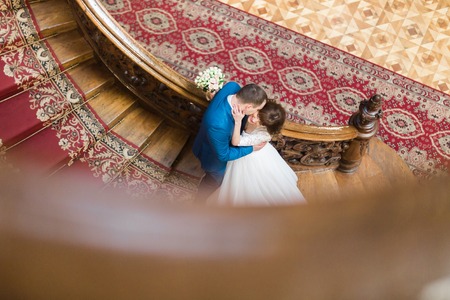 Handsome elegant groom kissing cheek of beautiful smiling bride on old wooden stairs at the background of rich interior.の写真素材