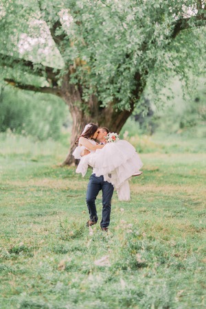 Young smiling groom carrying his beautiful wife in spring green park.の写真素材