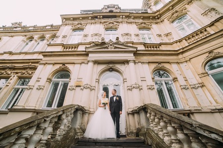Beautiful bride in white veil and handsome groom standing on the stairs against the backdrop of an old buildingの写真素材