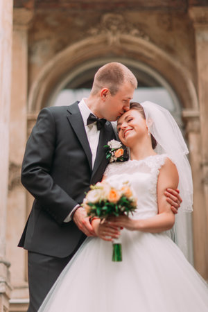Close-up photo of happy groom kissing bride's forehead on background old building.の写真素材