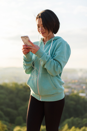 Sensual young asian woman listens to music in headphones during morning workout. Wonderful blue sky background.の写真素材