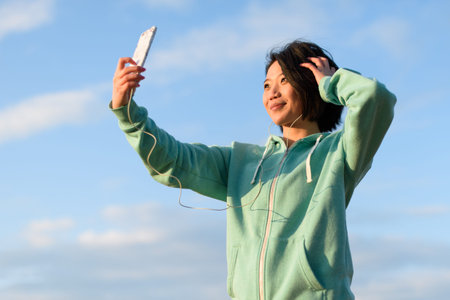 Sensual japanese woman with short hair taking selfie outdoor using her phone. Blue cloudy sky...