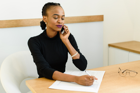 Serious confident young African or black American business woman on phone taking notes in office.の写真素材