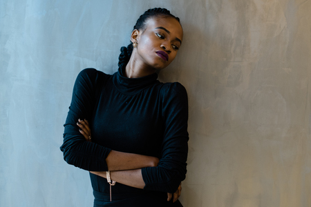 African-American sad woman dressed in black leaned against the wall. A lot of free space on a gray background.の写真素材