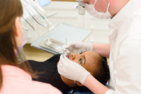 Young African-American ethnic black female opening her mouth while dentist in white latex gloves and mask and his assistant check condition of her teeth, close-upの写真素材