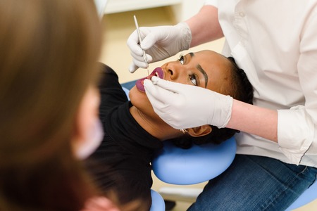 Young African-American ethnic black female opening her mouth while dentist in white latex gloves and mask and his assistant check condition of her teeth, close-upの写真素材